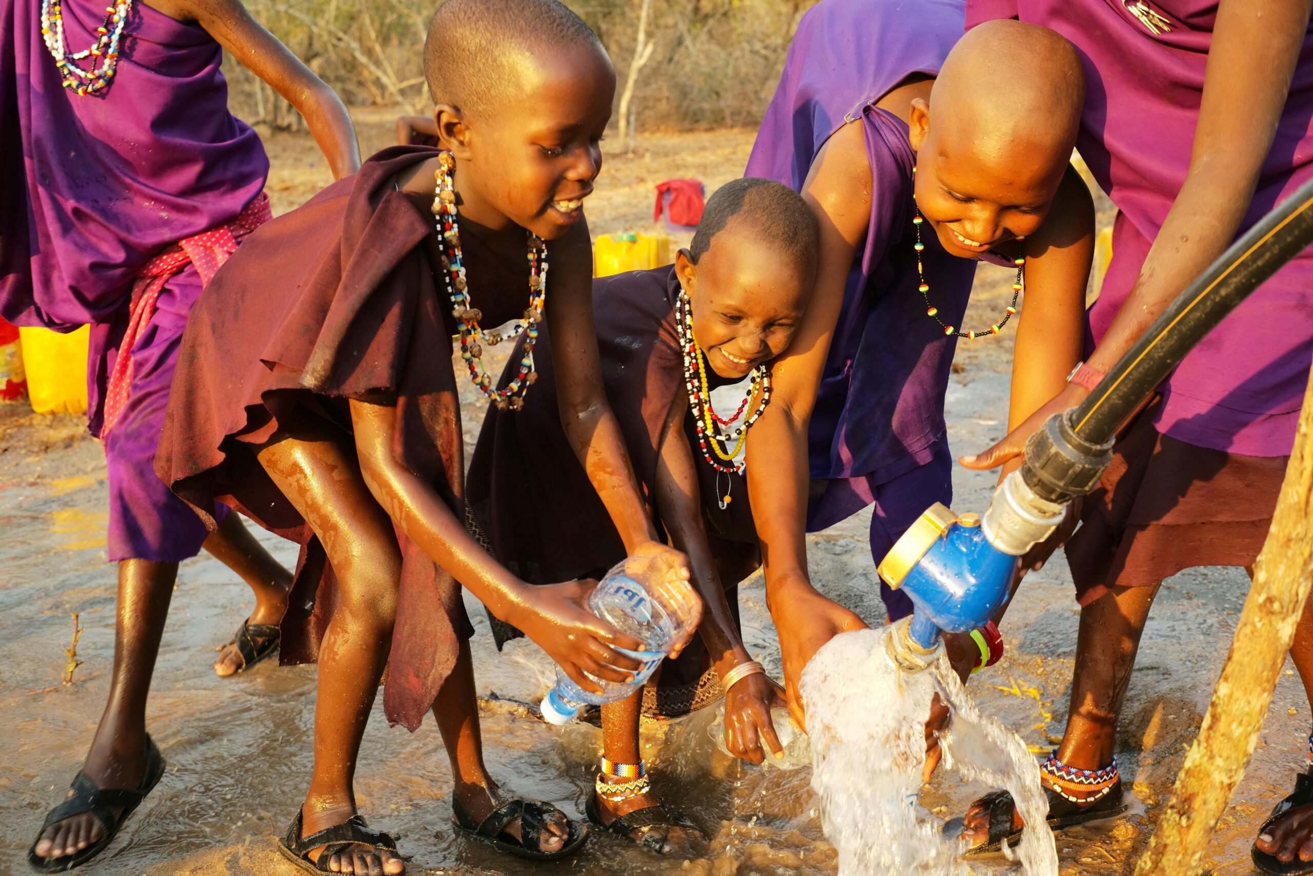 Smiling African children in traditional clothing enjoying water from a communal tap.